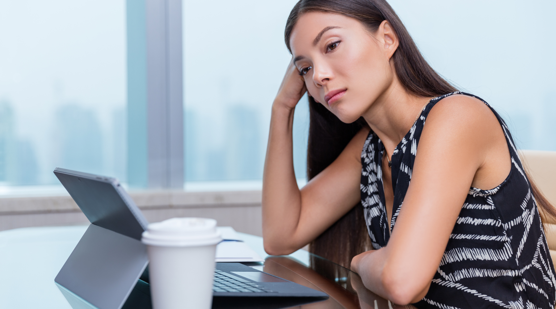 A pensive woman with long dark hair resting her head on her hand while looking at a laptop on a glass table with a coffee cup in the foreground, suggesting contemplation or distraction.