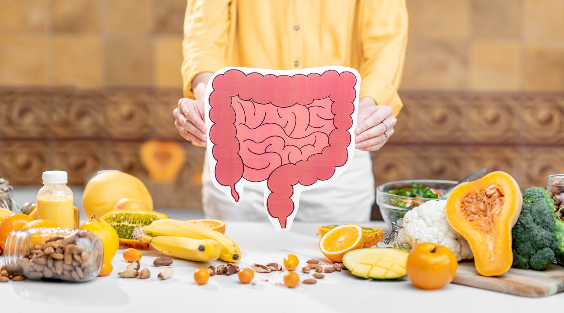Person holding an illustration of human stomach above a table with assorted fruits, nuts, and vegetables including bananas, oranges, mango, broccoli, cauliflower, and nuts.